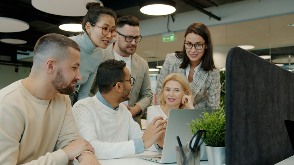 A group of colleagues gathered around a laptop, working together in an open-plan office
