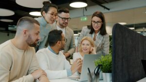 A group of colleagues gathered around a laptop, working together in an open-plan office